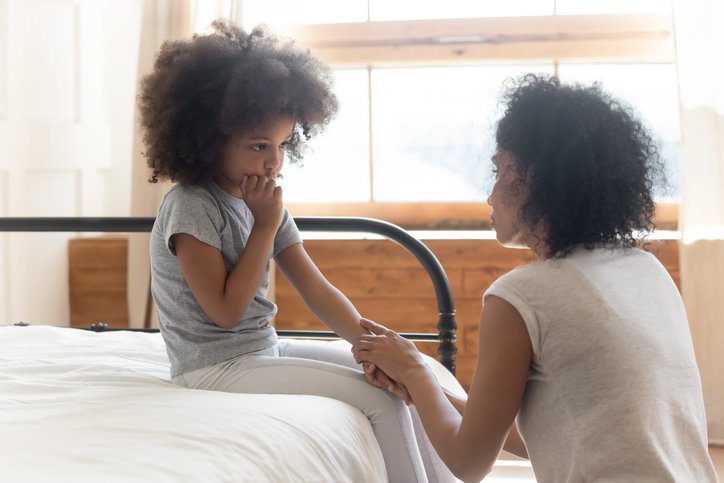 woman kneeling by child who is sitting on a bed, holding her hand and comforting her
